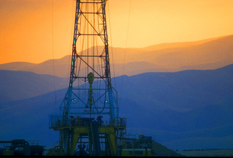 Drilling rig silhouetted against an orange sunset over hills